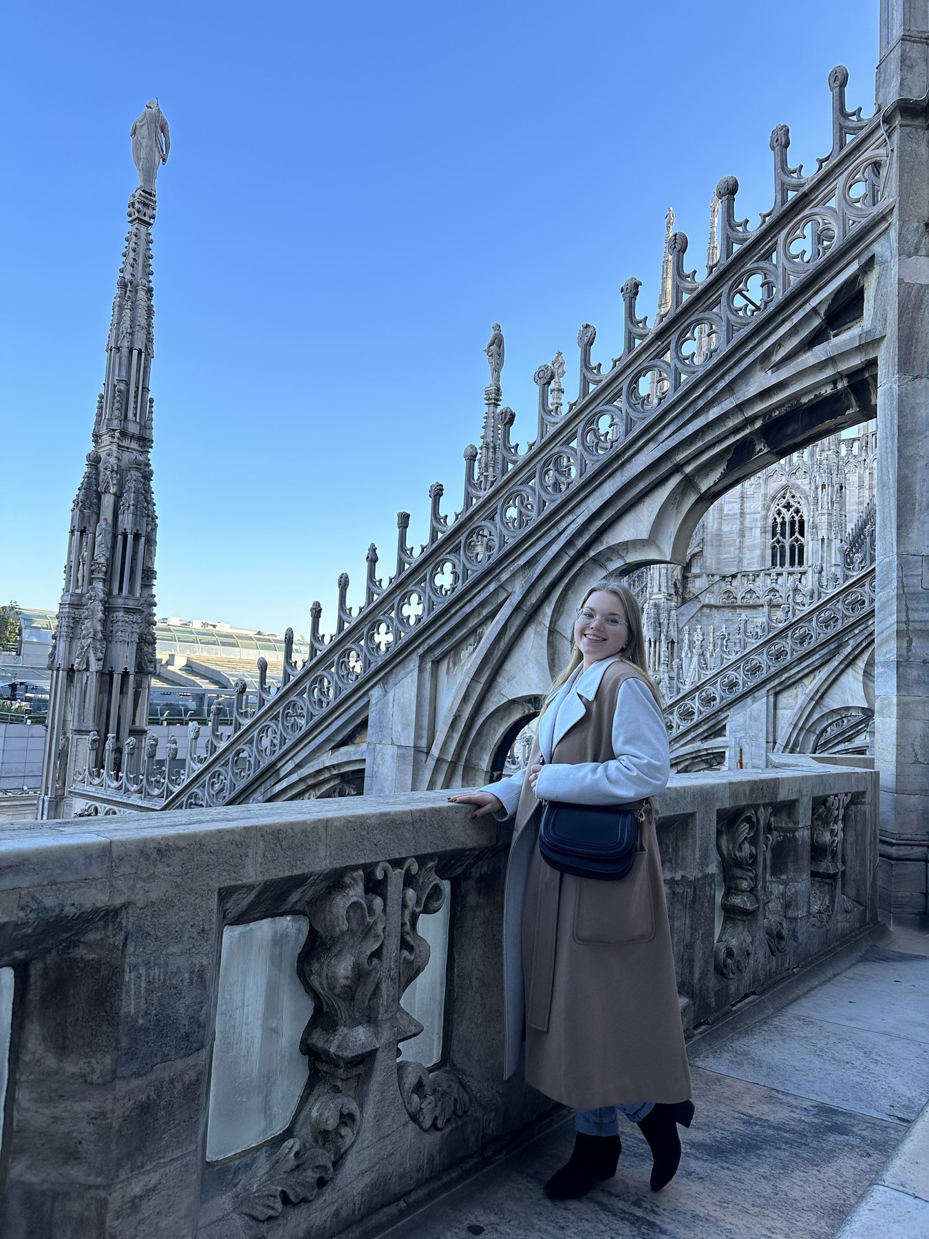 Una donna in cappotto sorride su un balcone con decorazioni in pietra intagliata in una cattedrale storica, sotto un cielo azzurro e limpido.